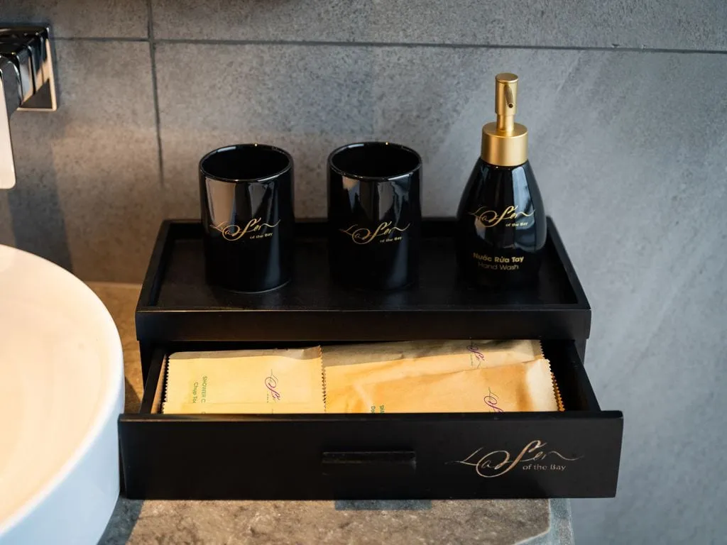 A black bathroom amenity tray with two cups, a hand wash dispenser and an open drawer of toiletry packets, all marked with gold 'La Sen of the Bay' branding, beside a white sink against grey stone tiles.