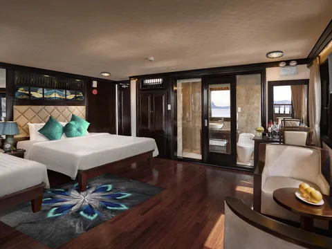 A luxury cruise cabin interior with two beds, teal cushions, dark wood floors, glass bathroom doors, seating area, and windows showing water and karst formations outside.