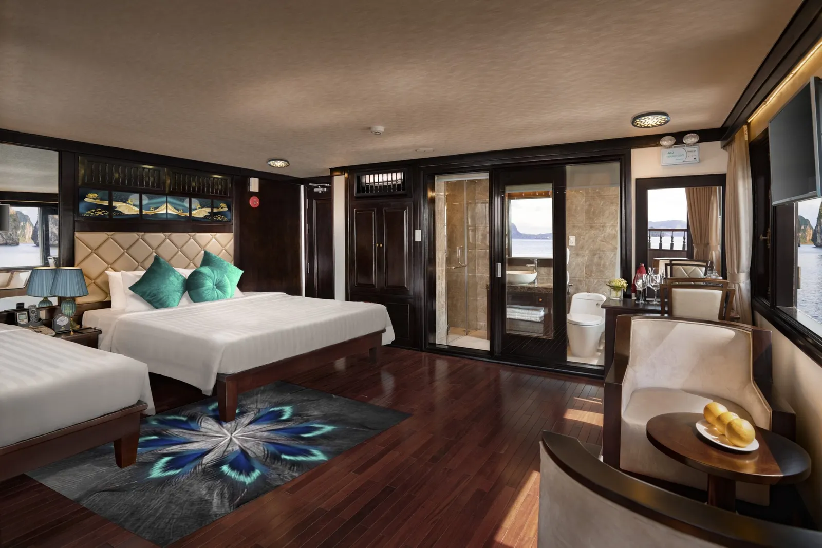 A luxury cruise cabin interior with two beds, teal cushions, dark wood floors, glass bathroom doors, seating area, and windows showing water and karst formations outside.
