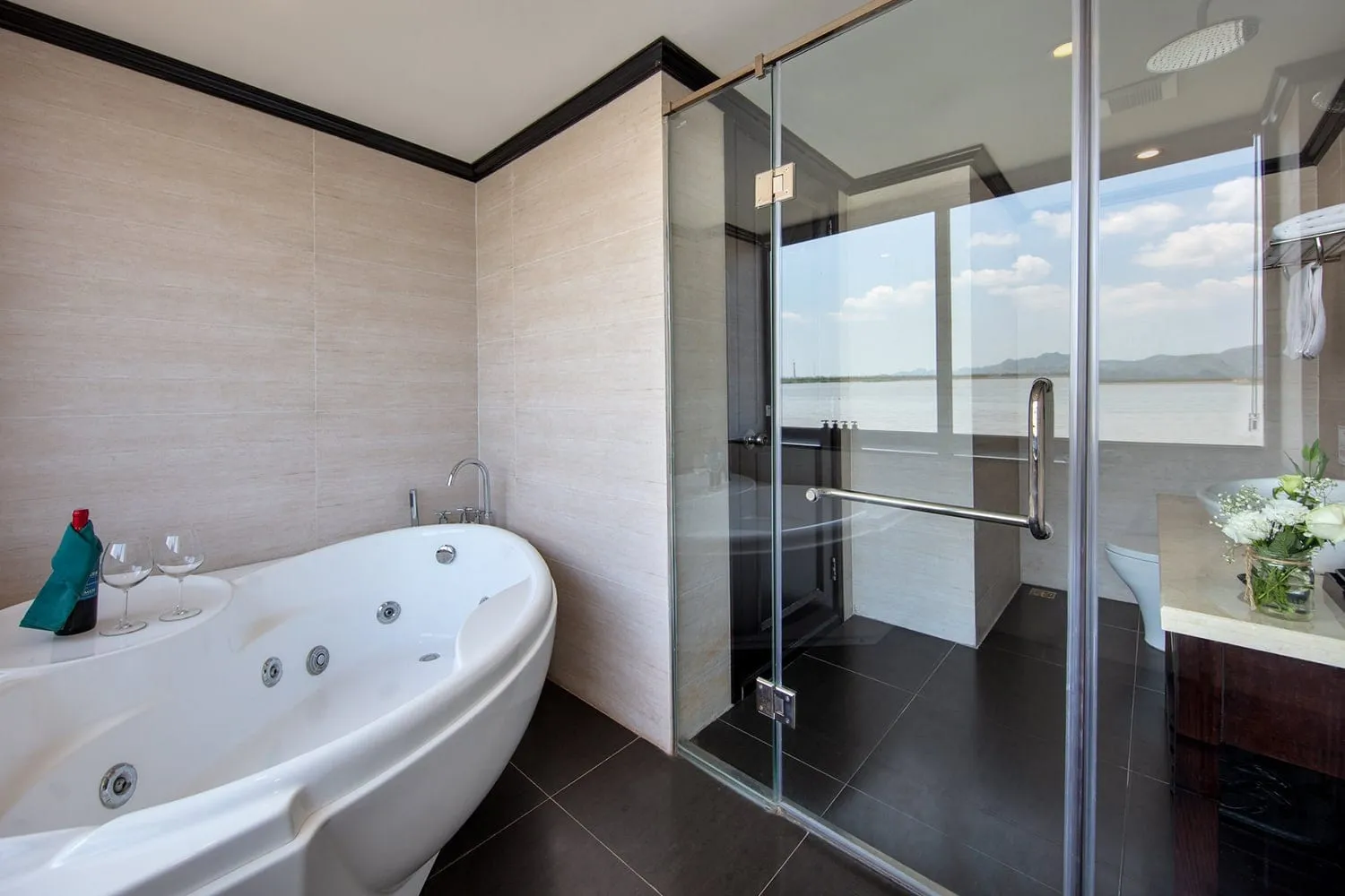 A bathroom with a white jacuzzi tub holding wine glasses and a bottle, a glass-enclosed shower, and a vanity with flowers, with a wide window showing flat water and distant hills.