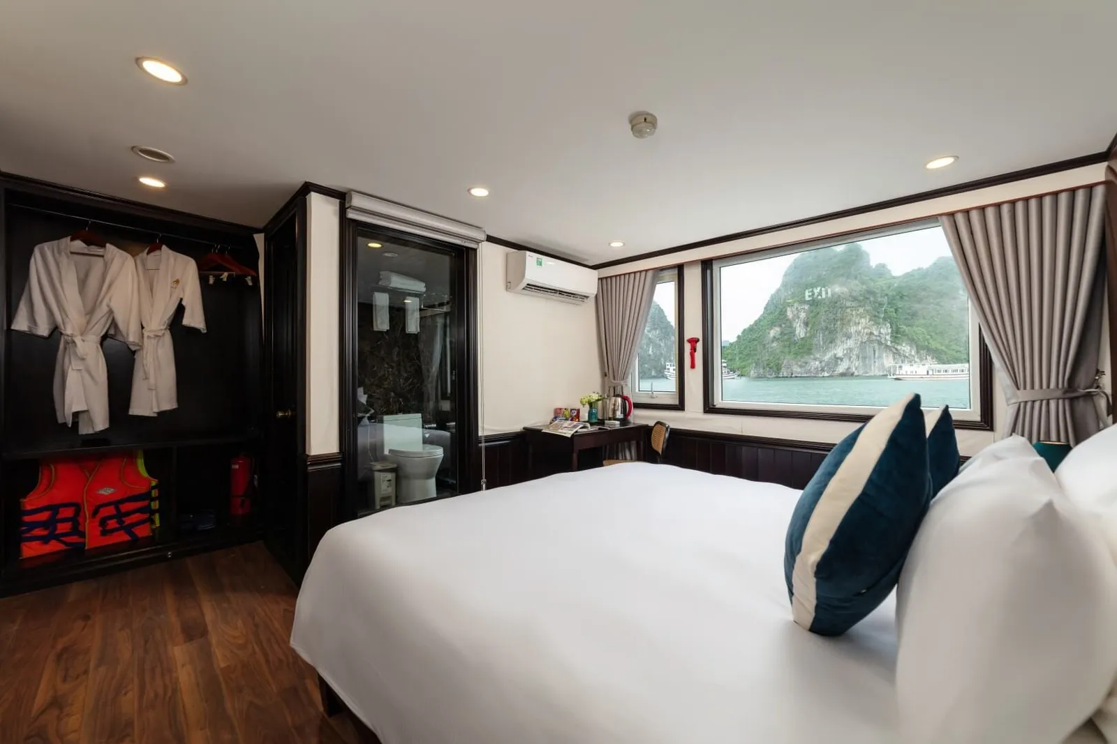 A cruise cabin with a white double bed, teal cushions, bathrobes and orange life jackets in an open wardrobe, a glass-walled bathroom, and a large window showing limestone karst islands on calm green water.