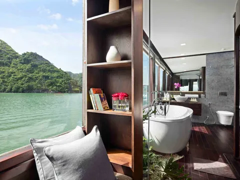 A luxury cabin interior with a freestanding bathtub, bathroom fixtures, wooden shelving, seating area, and large windows overlooking green karst mountains and calm bay water.