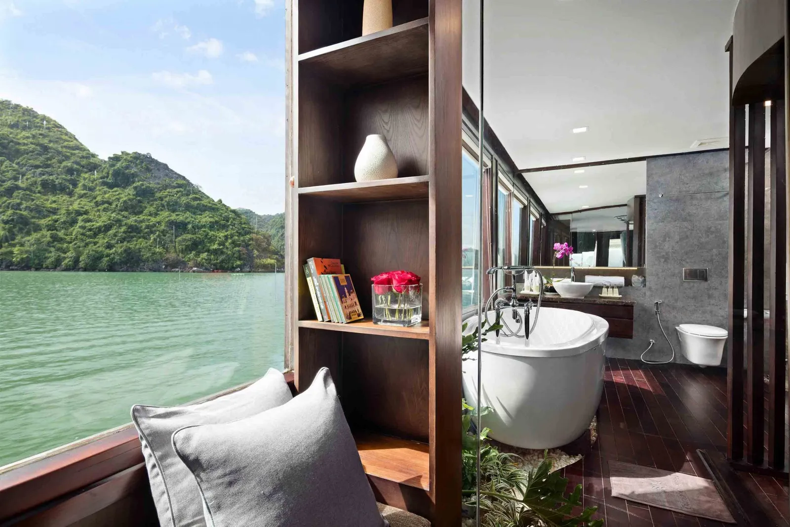 A luxury cabin interior with a freestanding bathtub, bathroom fixtures, wooden shelving, seating area, and large windows overlooking green karst mountains and calm bay water.