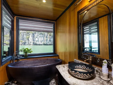 A wooden boat cabin bathroom with a dark bathtub and decorative sink, with windows showing limestone karst cliffs and green water outside.