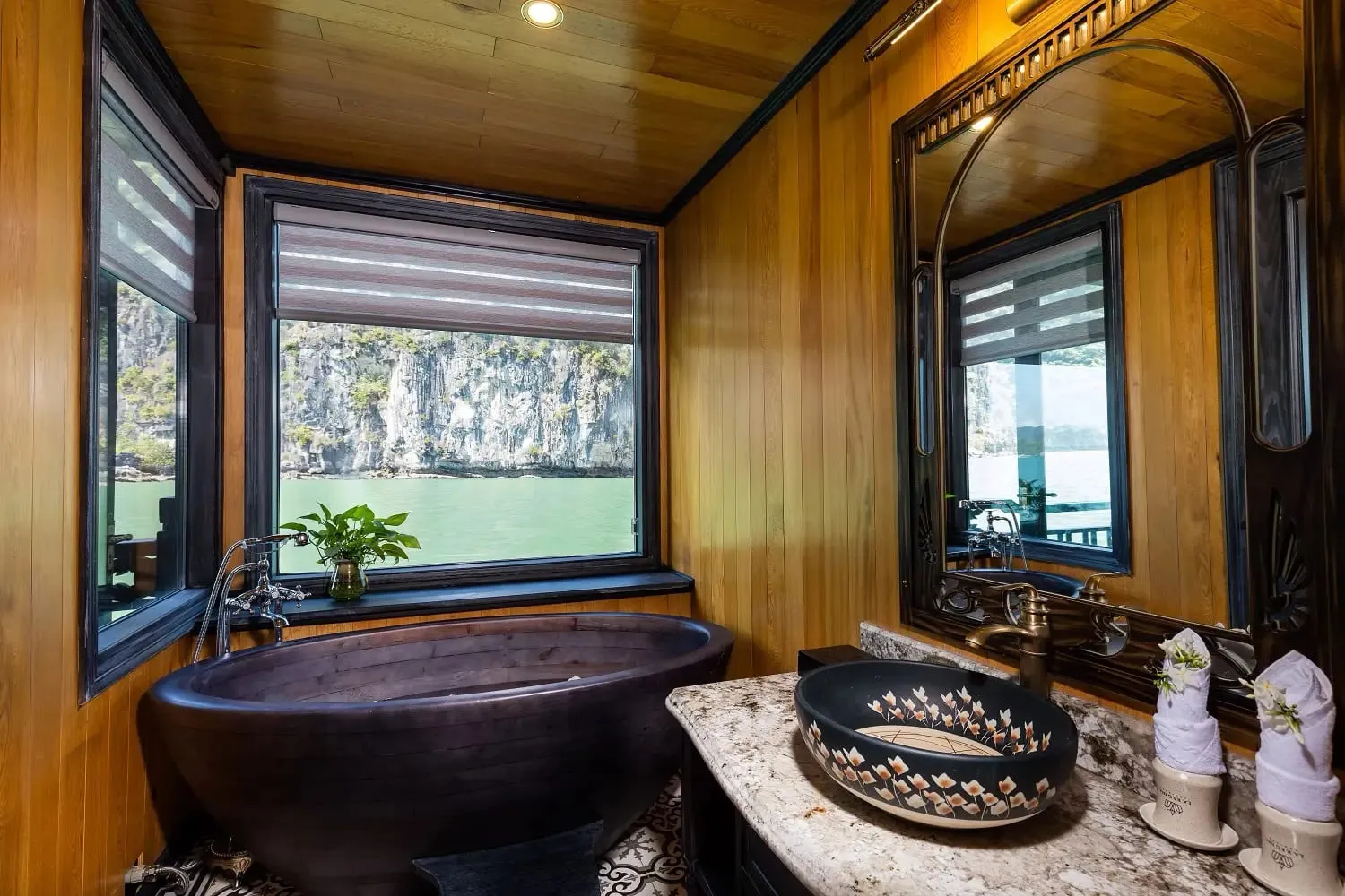A wooden boat cabin bathroom with a dark bathtub and decorative sink, with windows showing limestone karst cliffs and green water outside.