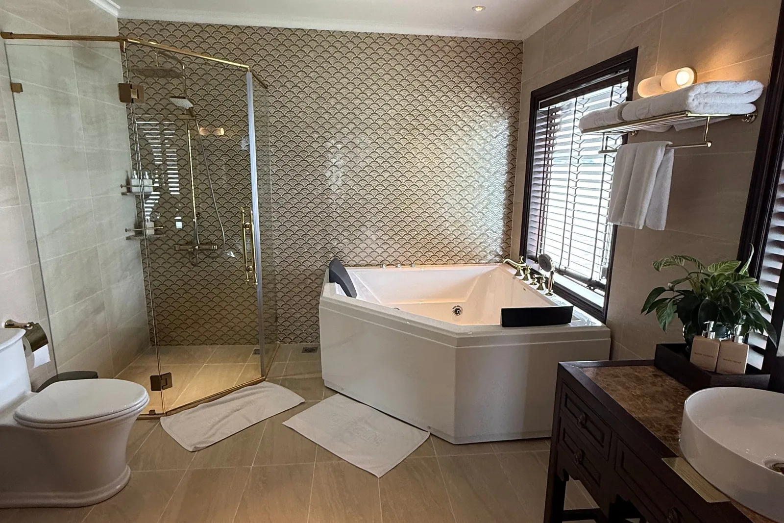 A spacious bathroom with a corner whirlpool tub, glass-enclosed shower with brass fittings, toilet, wooden vanity with a vessel sink, towels on a rack, and a window with wooden blinds.