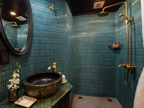 A bathroom with teal vertical tiles, a walk-in shower with brass fixtures, a dark ornate vessel sink on a green marble vanity, an oval black-framed mirror and a white orchid in a vase.