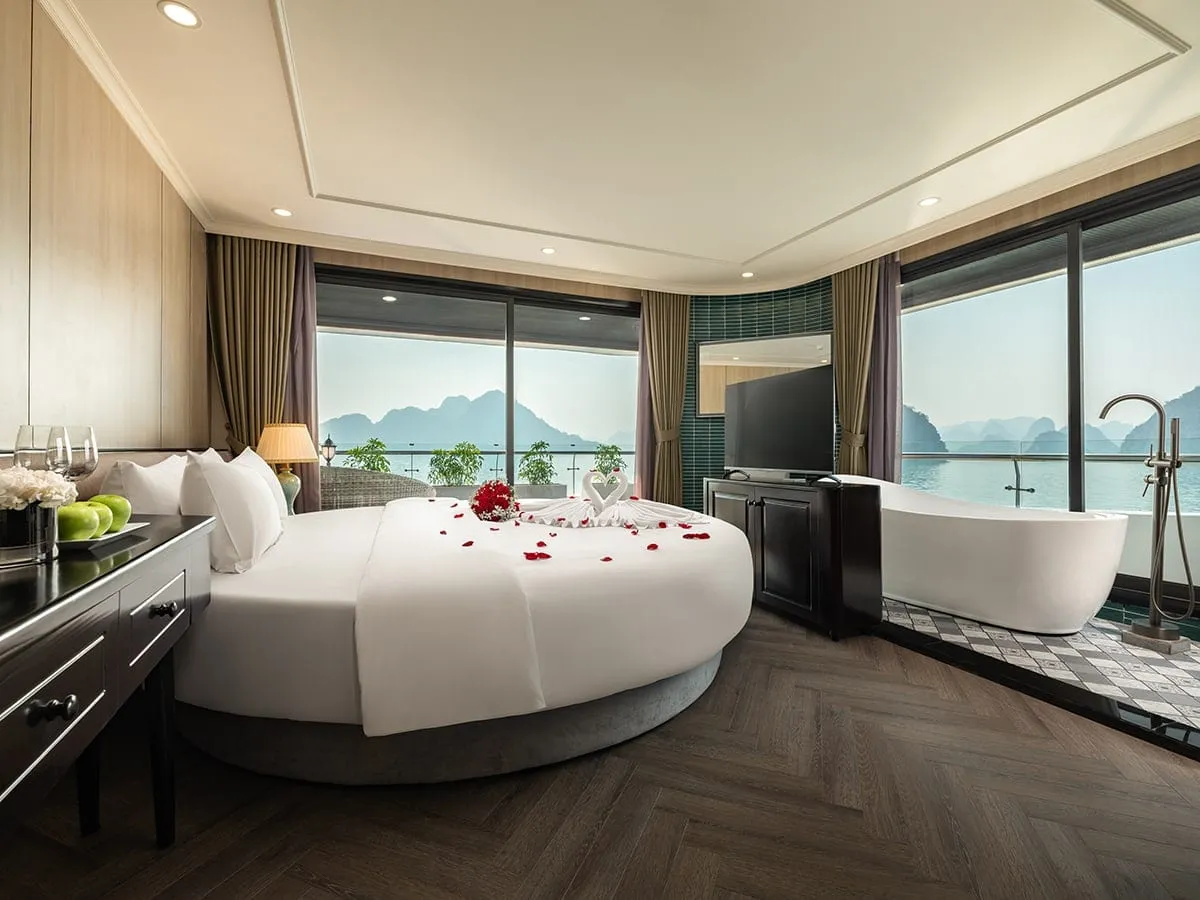 A cabin with a round bed decorated with rose petals and a towel swan, a freestanding bathtub, and a TV, with floor-to-ceiling windows overlooking calm water and limestone karsts.