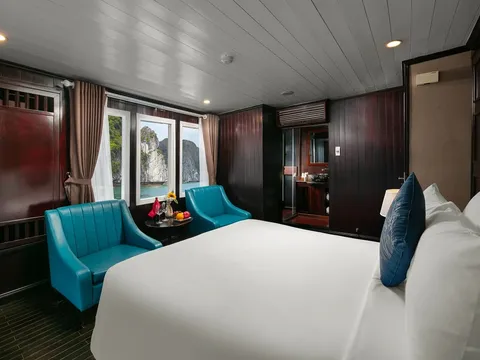 A dark-wood cruise cabin with a white double bed, blue pillow, two teal armchairs and a small table with fruit, with windows showing limestone karst cliffs over green water and an en-suite bathroom behind.