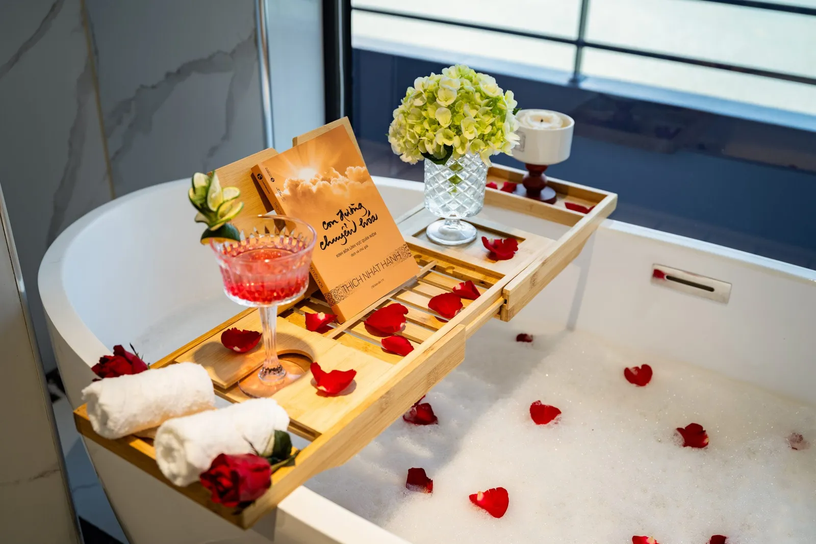 A freestanding bathtub full of foam and red rose petals with a bamboo tray holding a pink cocktail, a Vietnamese book, flowers, a candle and rolled towels, beside a large window.