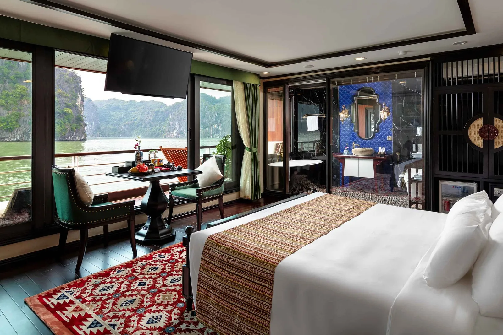 A luxury cruise cabin with large windows overlooking Ha Long Bay's limestone karsts, featuring a bed, seating area, and ensuite bathroom with blue tile.