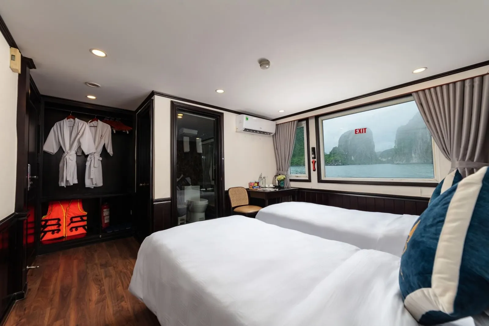 A twin-bed cabin with white linens, bathrobes and orange life jackets in an open wardrobe, a glass-door bathroom, and a large window showing misty limestone karst islands on the water, with a small gold emblem on the blue cushion and a red EXIT sign visible through the window.