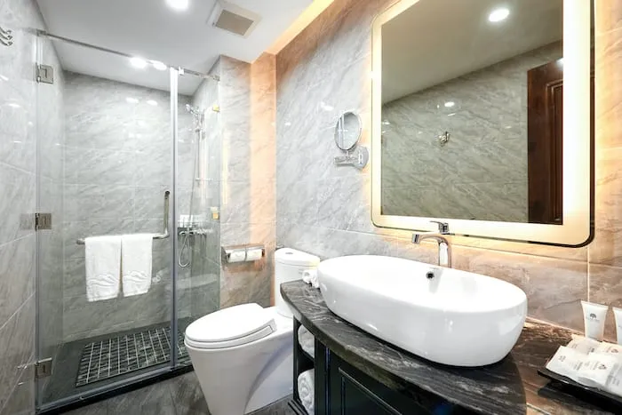 A modern bathroom with marble walls, glass shower enclosure, white vessel sink, backlit mirror, and toilet.