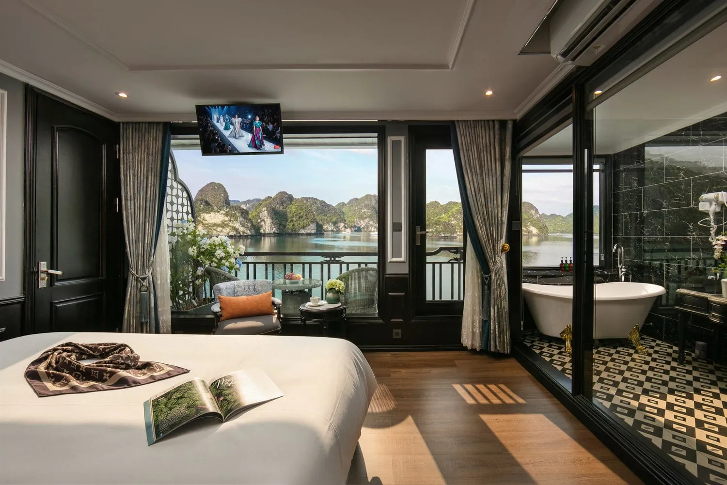 A luxury boat cabin with a bed, balcony seating, and ensuite bathroom overlooking limestone karsts and calm water.