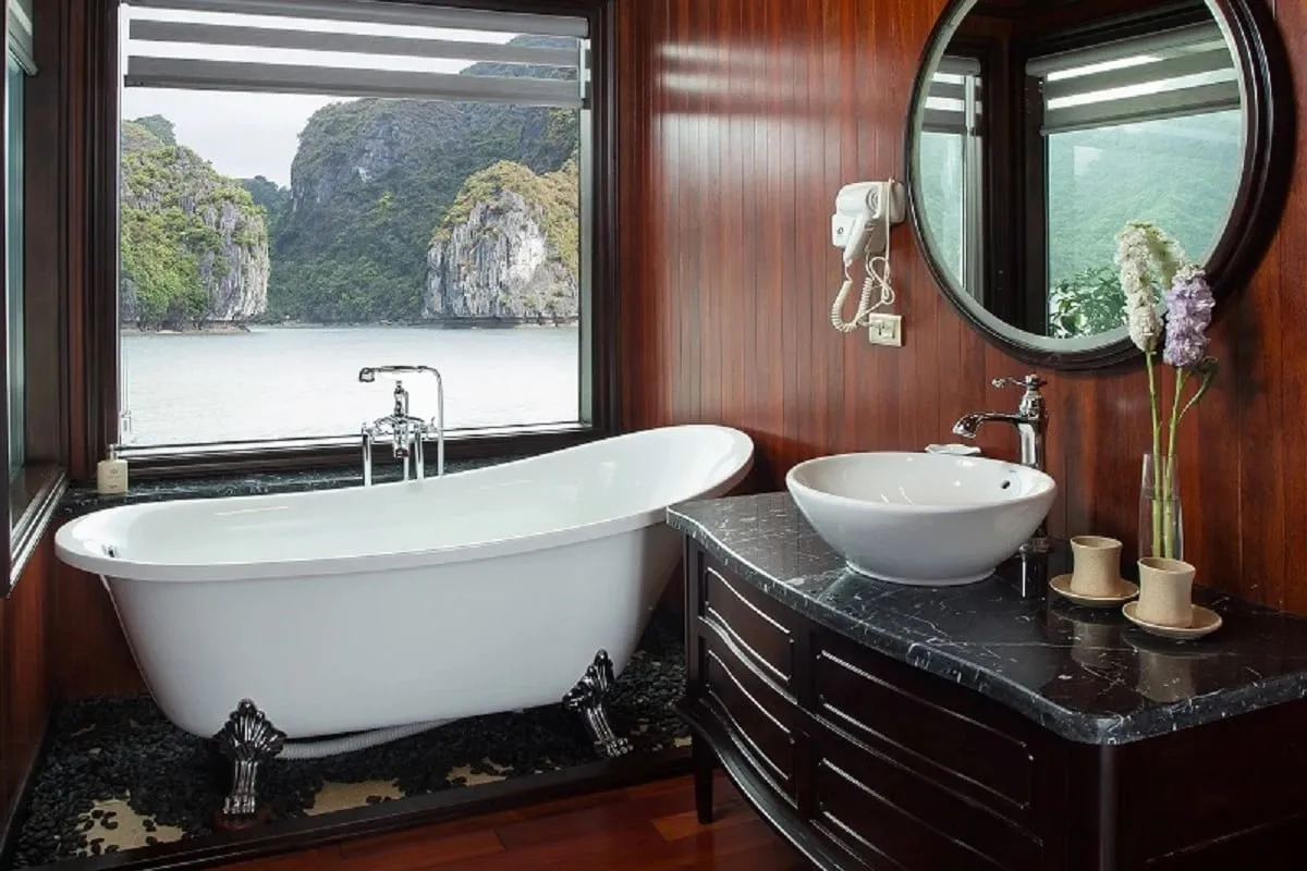 A luxury bathroom cabin with a freestanding bathtub and sink facing a window that shows limestone karsts rising from calm water.