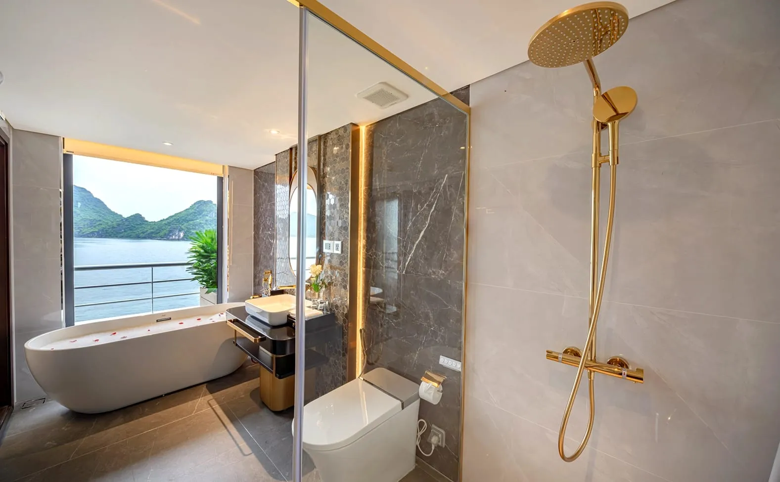A luxury boat bathroom with gold fixtures, freestanding tub, glass shower, and large window showing karst mountains and bay water.