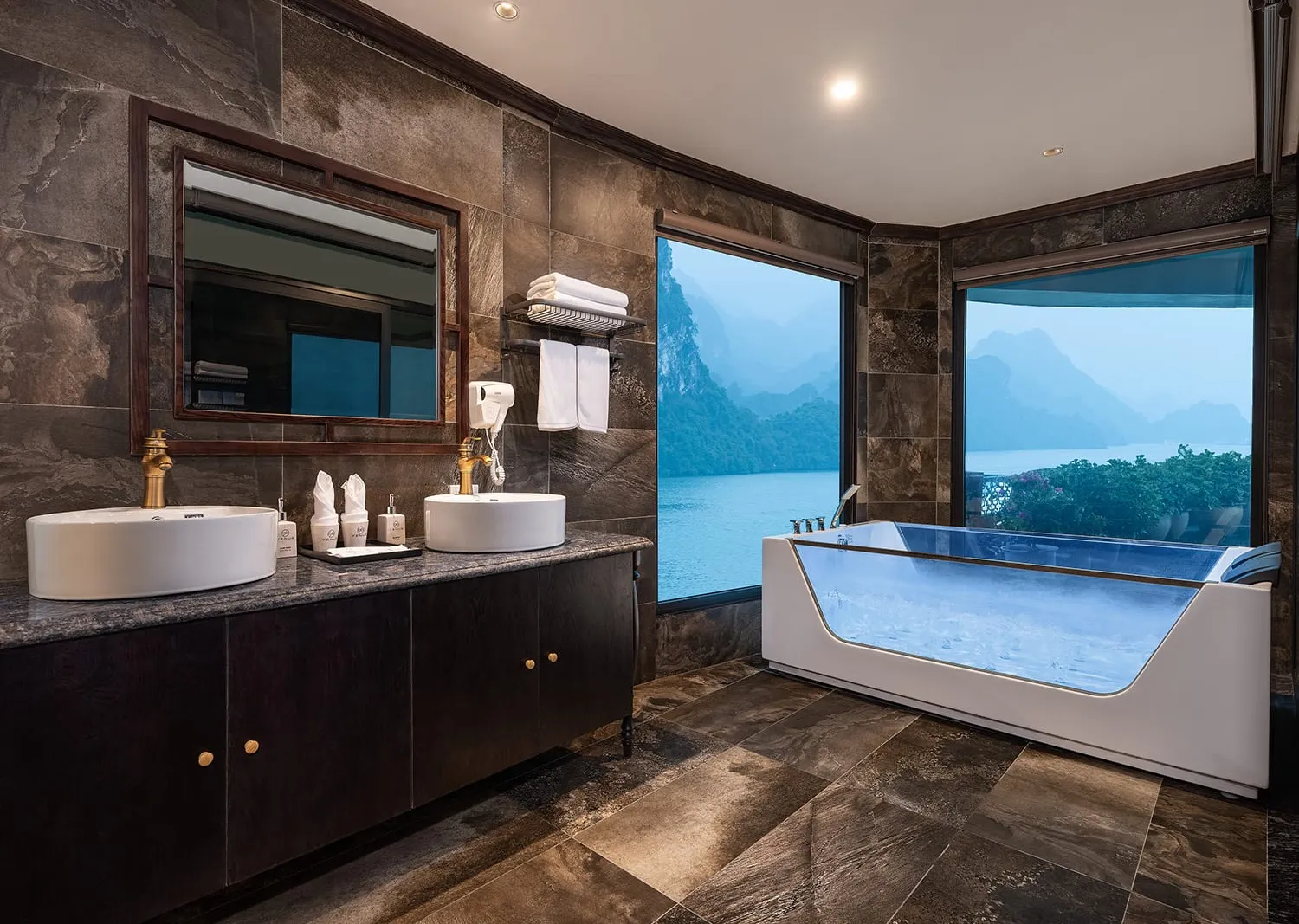 A luxury bathroom with dark stone tiles, double vessel sinks, a glass-sided soaking tub, and large windows overlooking karst limestone islands on calm water.