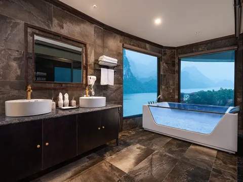 A luxury bathroom with dark stone tiles, double vessel sinks, a glass-sided soaking tub, and large windows overlooking karst limestone islands on calm water.