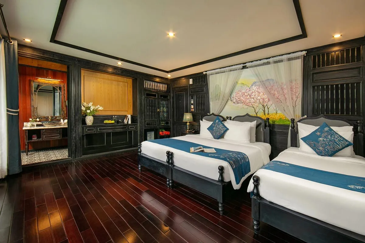 A cruise boat cabin with two beds, dark wood furniture, an en-suite bathroom visible through a doorway, and a decorative wall mural behind the beds.