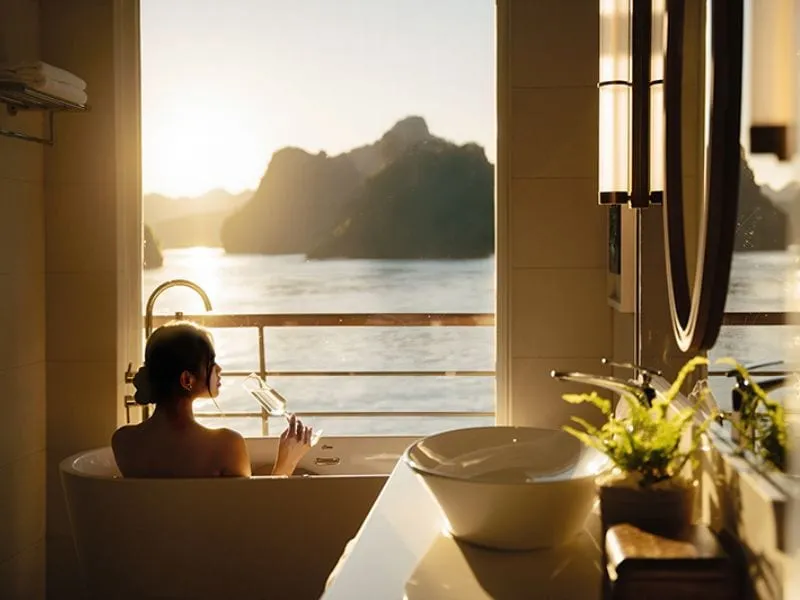 A person relaxes in a bathtub inside a boat cabin with a large window overlooking dramatic limestone karsts and calm water at golden hour.