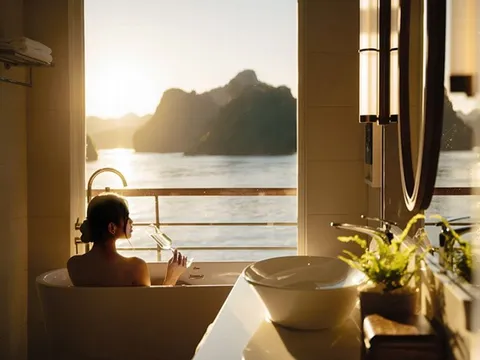 A person relaxes in a bathtub inside a boat cabin with a large window overlooking dramatic limestone karsts and calm water at golden hour.