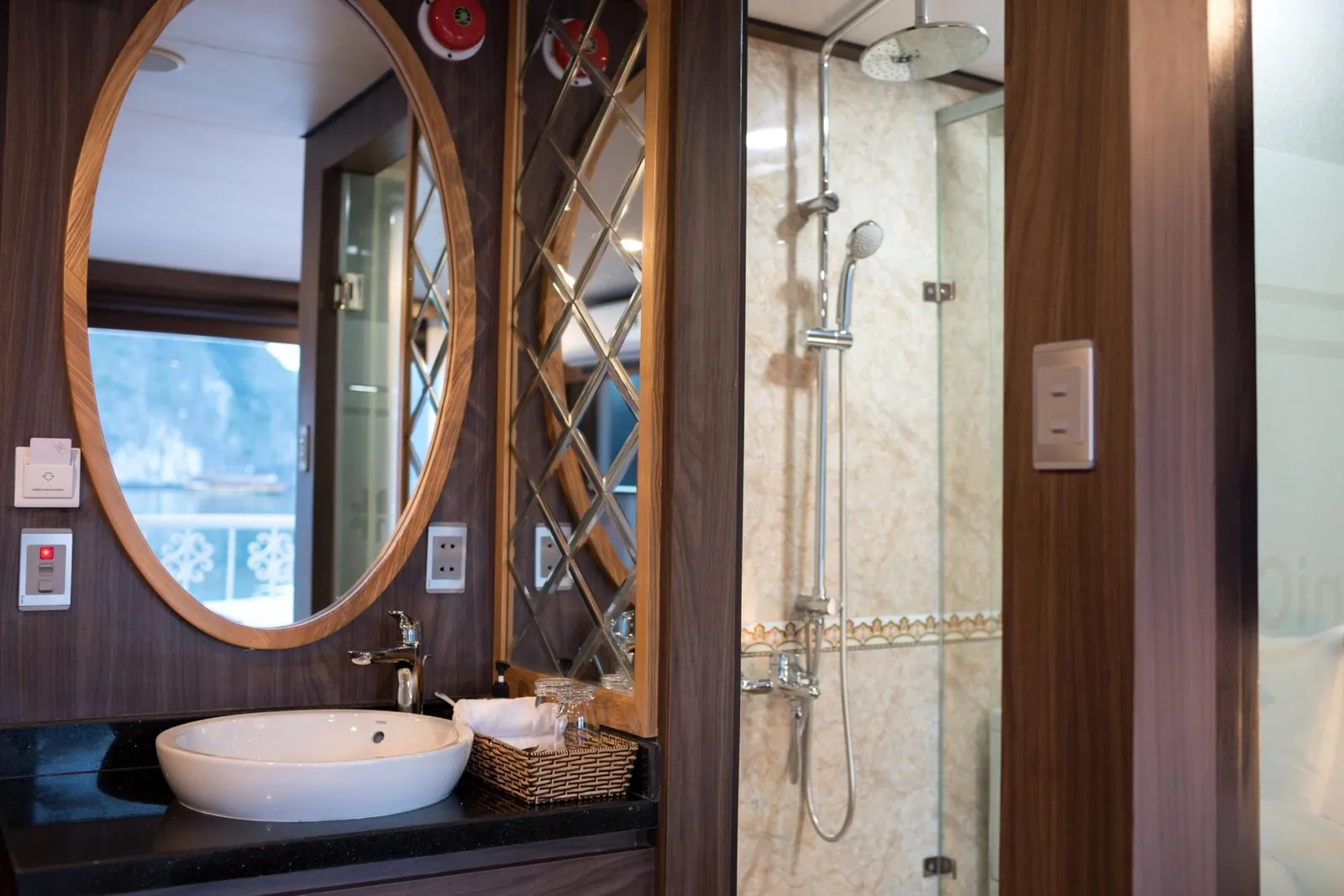 A cabin bathroom with a white vessel sink, oval wood-framed mirror, and glass-enclosed shower, with a mirror reflecting a window that looks out on misty limestone karst and water.