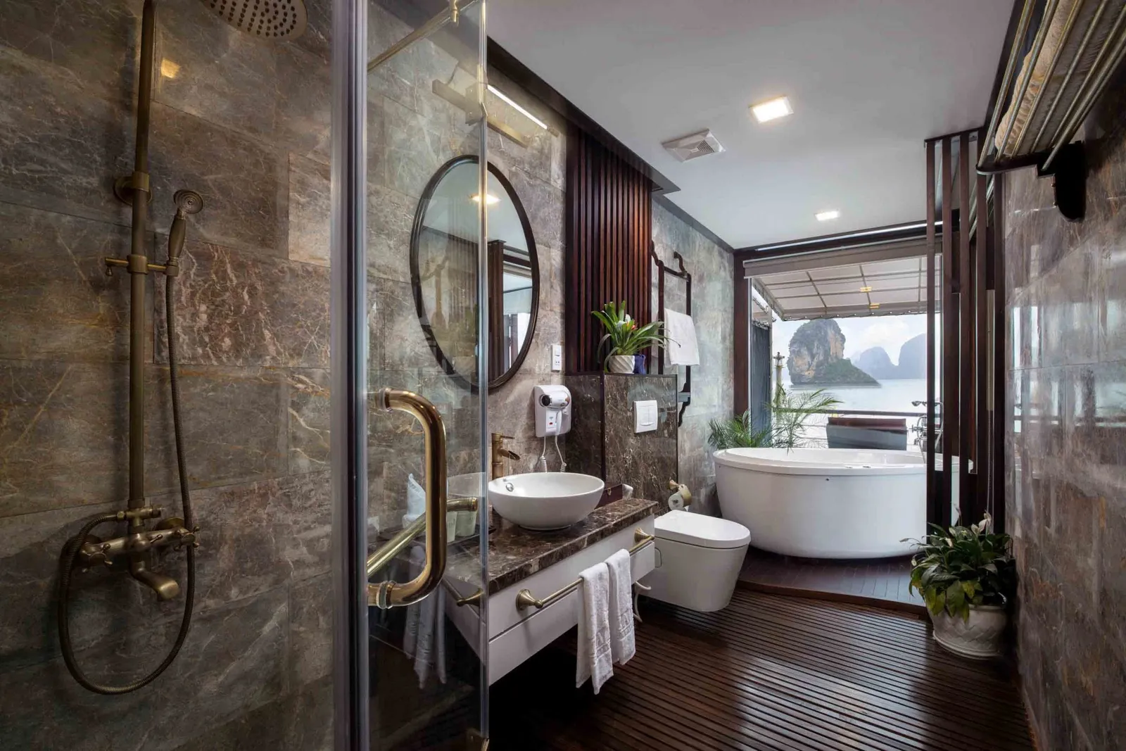 A luxury boat bathroom with marble walls, a freestanding tub, and a view of limestone karsts through the open door.