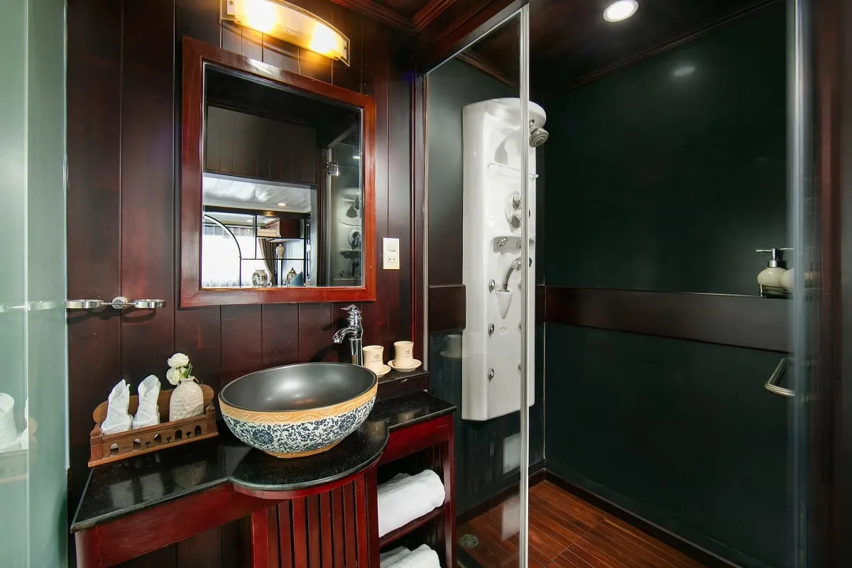 A dark wood-paneled bathroom with a blue-and-white ceramic vessel sink, a mirror, a glass-enclosed shower with a white jet panel, and folded towels on a shelf.