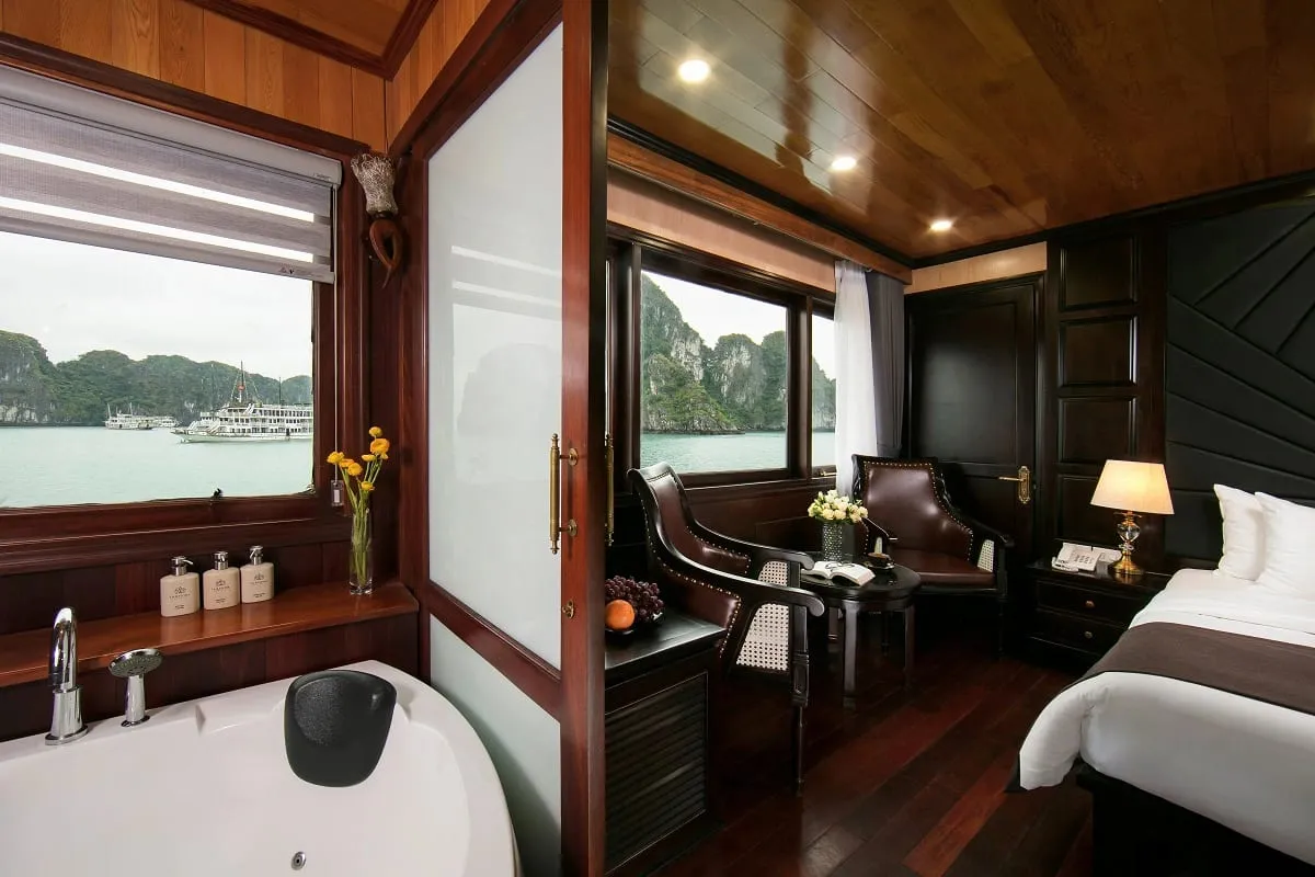 A luxury cabin interior with bathtub, bed, and seating area with windows showing limestone karsts and cruise boats on calm water.