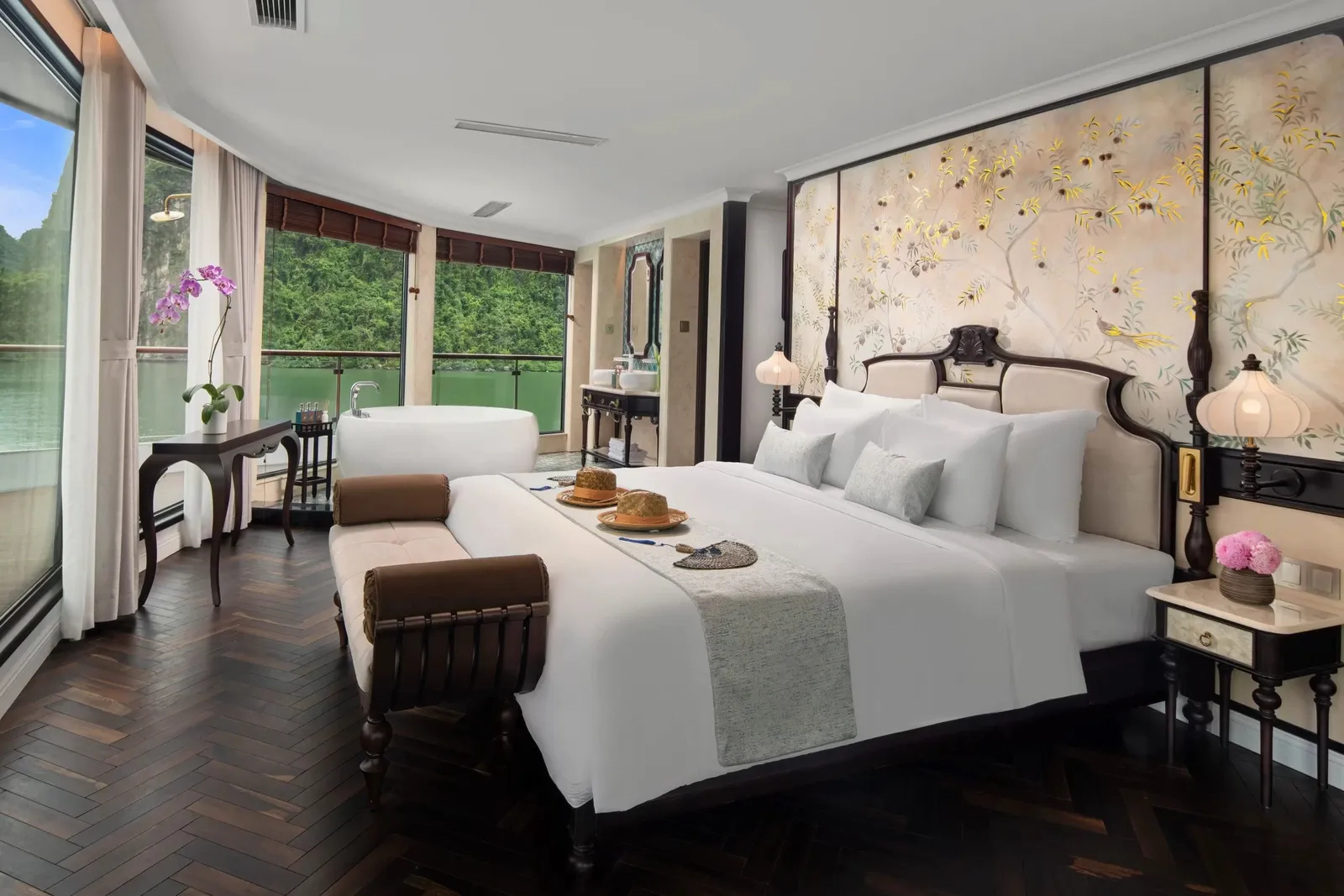 A luxury cabin interior with a king bed, freestanding bathtub, decorative floral wall panels, and floor-to-ceiling windows overlooking green water and karst limestone formations.
