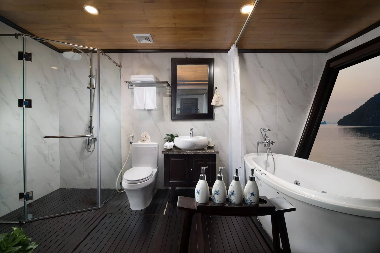 A marble-tiled en-suite bathroom with a glass shower, toilet, vanity with mirror, and freestanding bathtub, with a slanted window showing calm water and a rocky karst island at dusk.