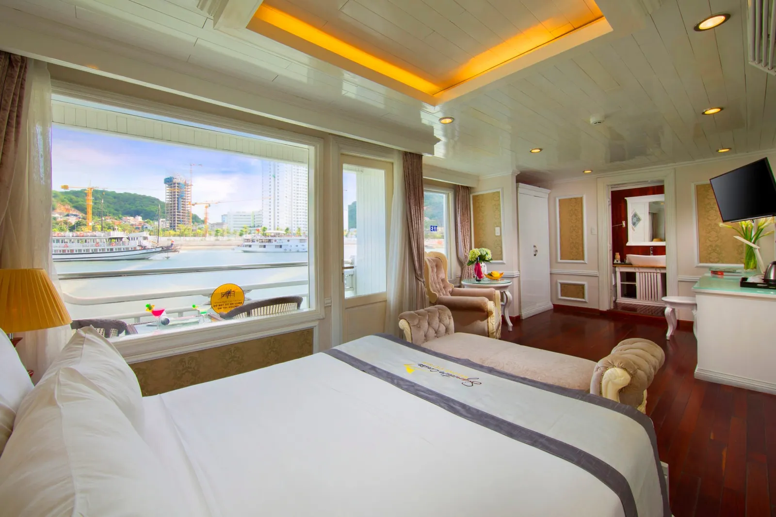 A cruise cabin with a large bed, a runner embroidered with a logo, armchairs, a TV and an en-suite bathroom, with a big window showing the harbour, other cruise ships and construction cranes.