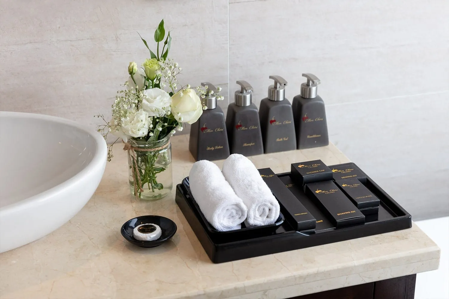 A bathroom vanity with a white vessel sink, a jar of white flowers, branded dispensers, rolled white towels and a tray of amenity boxes on a marble countertop.