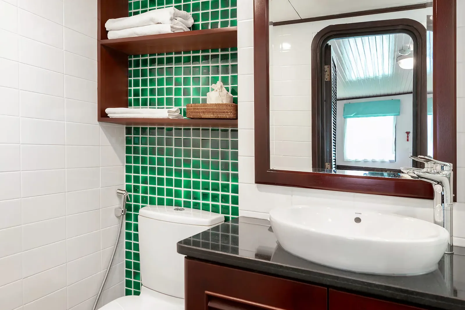 A cabin bathroom with white wall tiles, a green mosaic tile panel, dark wood shelves holding towels, a toilet, and a wooden louvered vanity with a white basin, with a mirror reflecting the cabin's arched door and window.