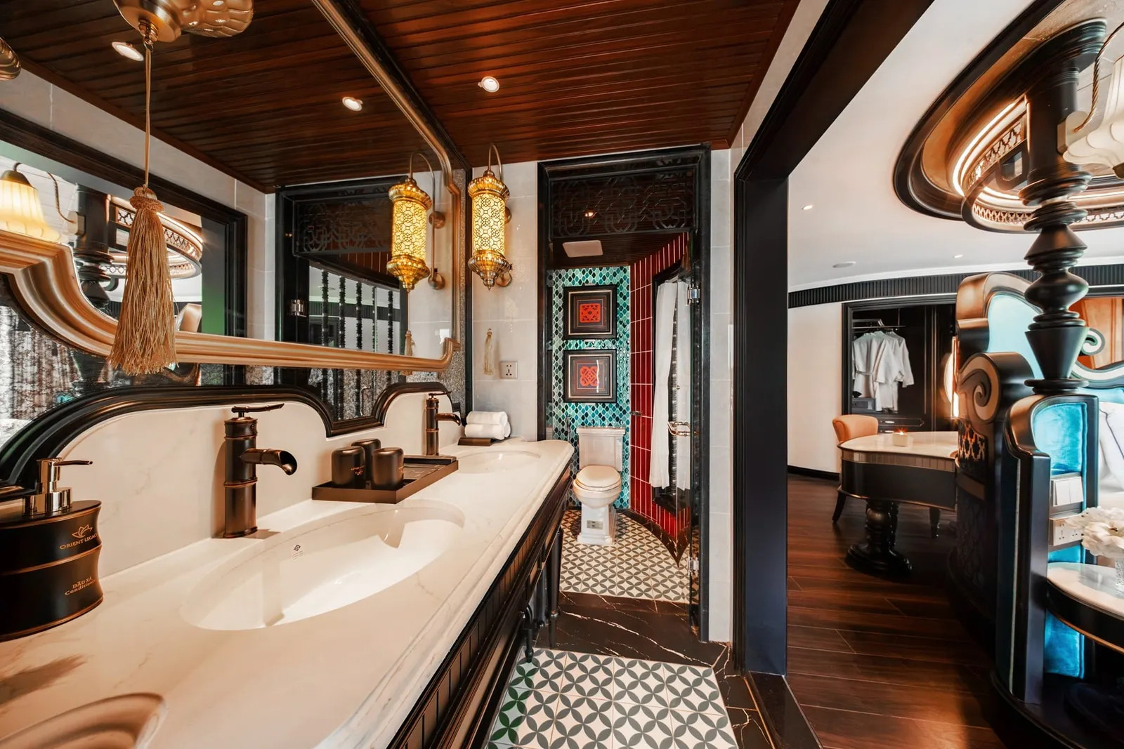 An ornate cabin bathroom with a double marble vanity, bronze taps, brass lanterns and patterned tiles, opening onto a bedroom with dark wood floors, and a small logo on the soap dispenser.
