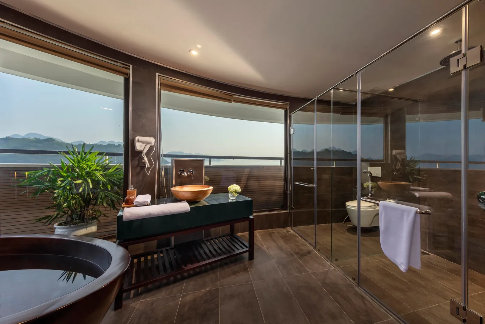 A bathroom with a dark freestanding tub, a copper vessel sink on a vanity, a glass-enclosed shower and toilet, and large windows looking out on hazy limestone hills and water.