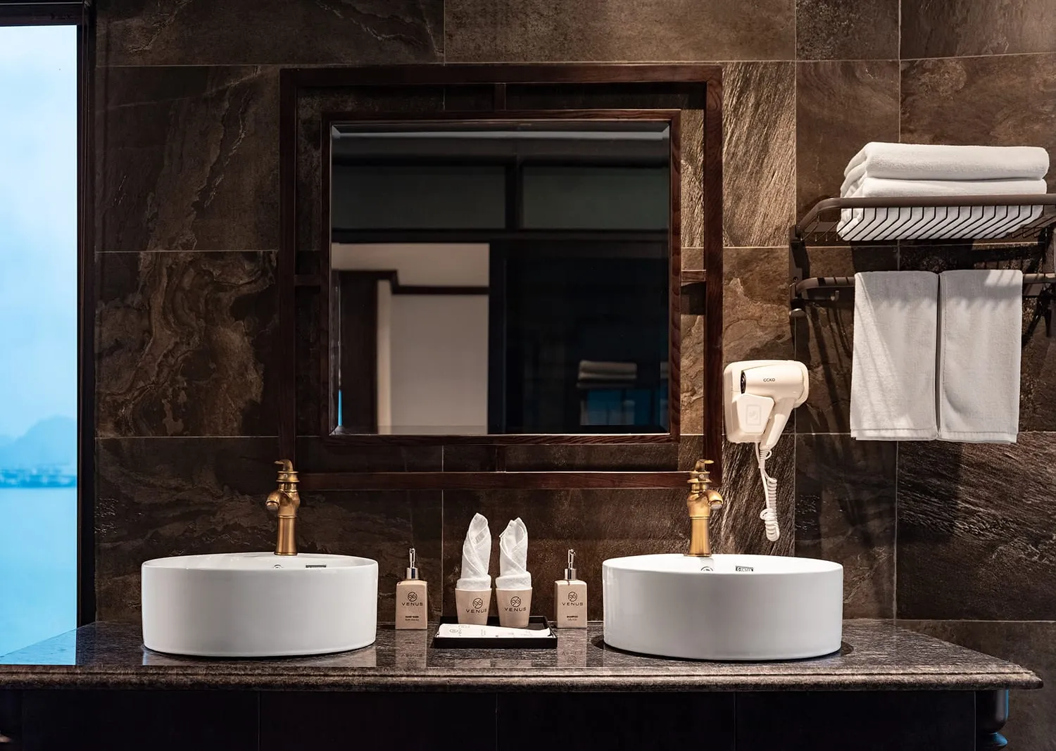 A double-sink bathroom with dark marble tiles, brass faucets, a wood-framed mirror, white towels, a wall-mounted hair dryer, and toiletry bottles labeled 'VENUS' on the counter; a window at left reveals calm water and karst hills outside.