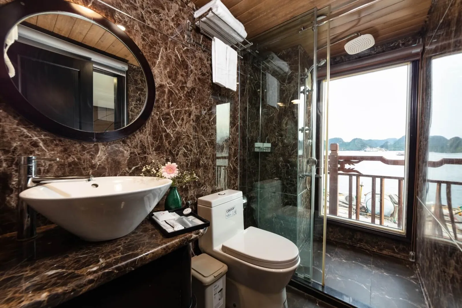 A marble-clad ensuite bathroom with a vessel sink, oval mirror, toilet and glass shower, with a window showing a wooden railing, calm water and limestone karst islands beyond.