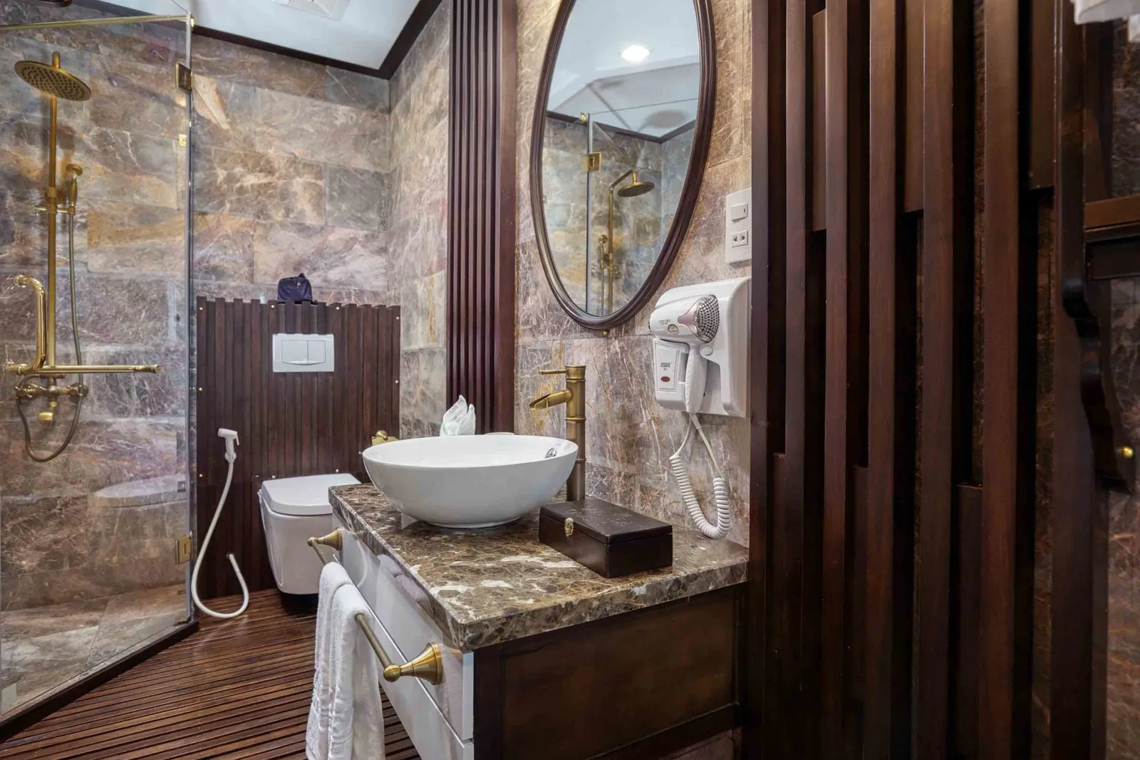 A bathroom with marble walls, dark wood slat paneling and flooring, a glass shower with brass fixtures, a wall-hung toilet, a vessel sink on a marble vanity, an oval mirror and a wall-mounted hair dryer.