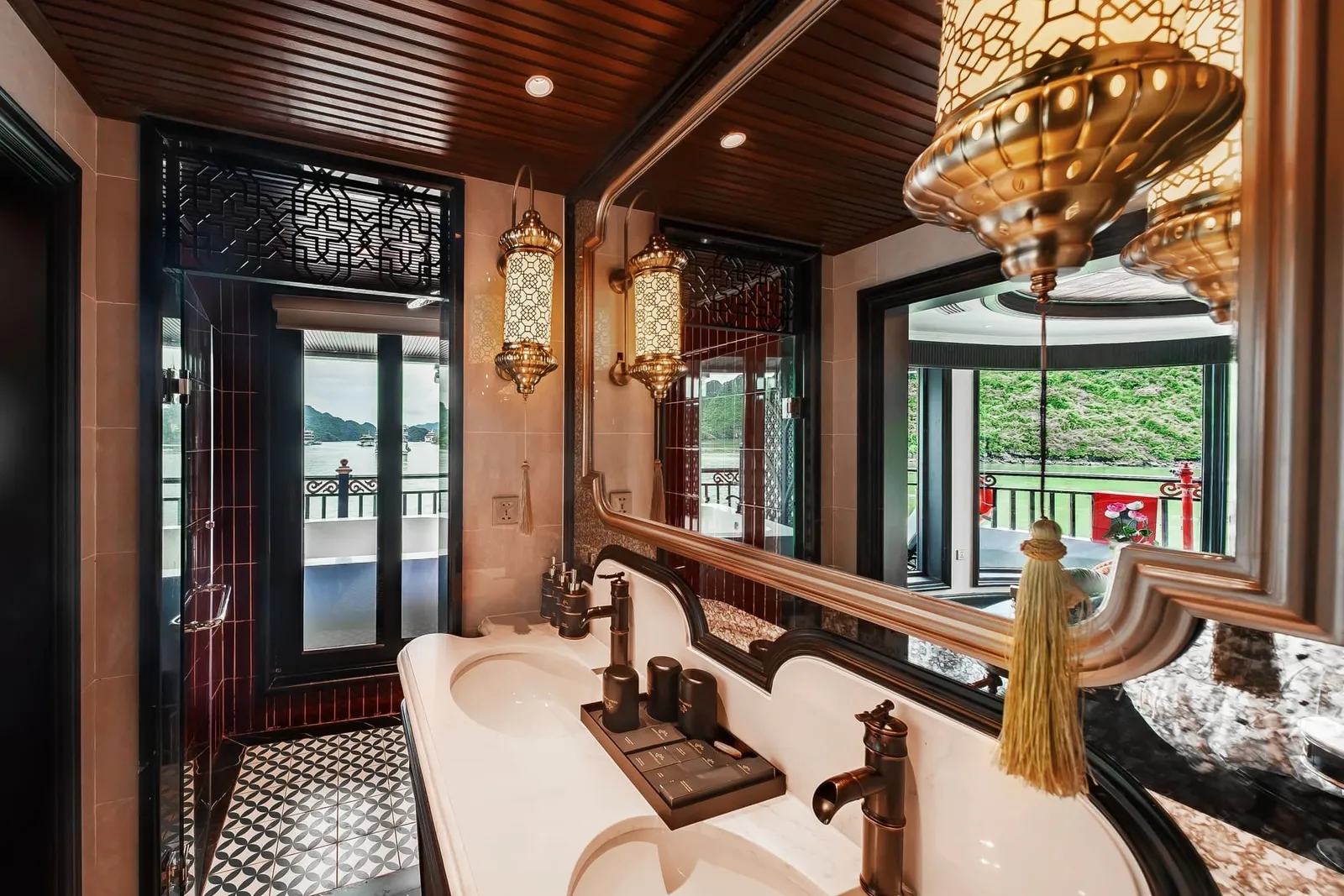 A cruise cabin bathroom with a marble double vanity, bronze taps, ornate lanterns, a large mirror and a glass door opening onto a balcony with a view of green karst hills and calm water.