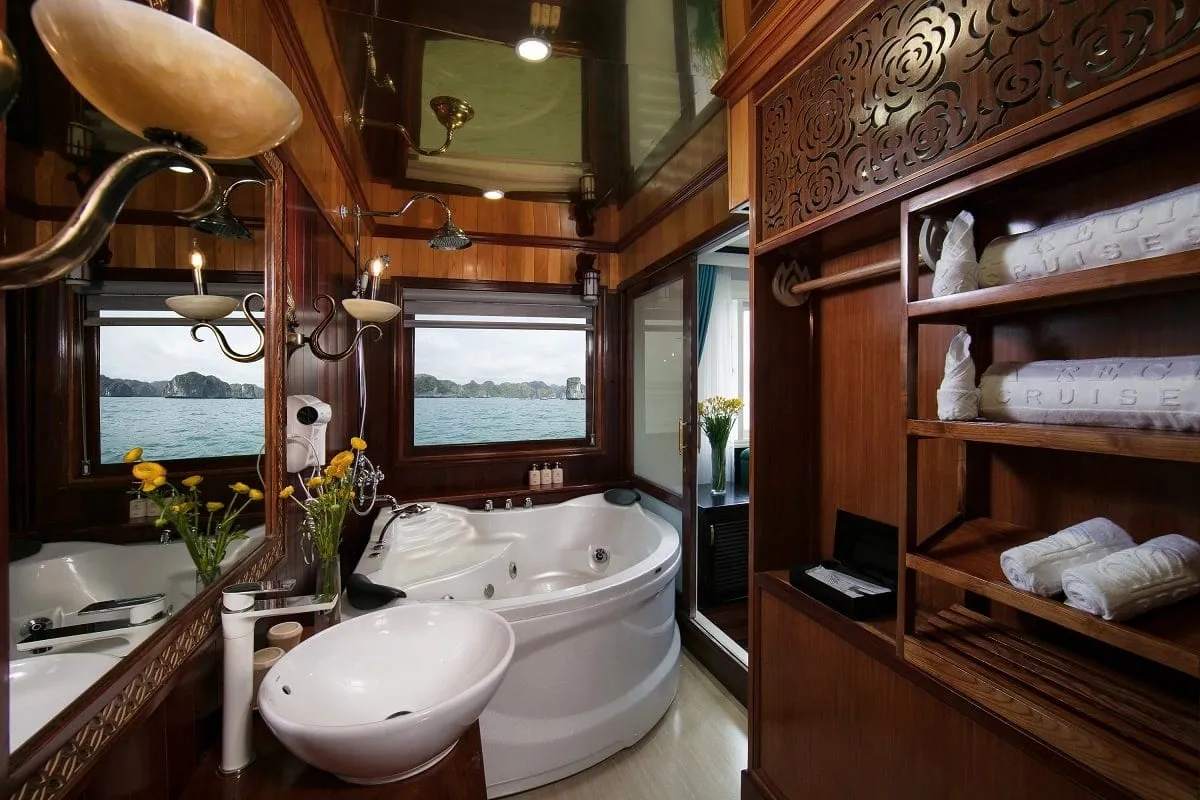 A luxury cruise cabin bathroom with carved wood finishes, dual white bathtubs, yellow flowers, and windows showing limestone karsts in the bay.