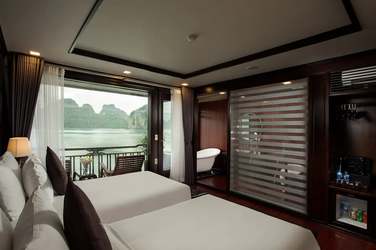 A cruise boat cabin with two beds, dark wood paneling, a glass-enclosed bathroom, and a window showing karst limestone mountains and water outside.