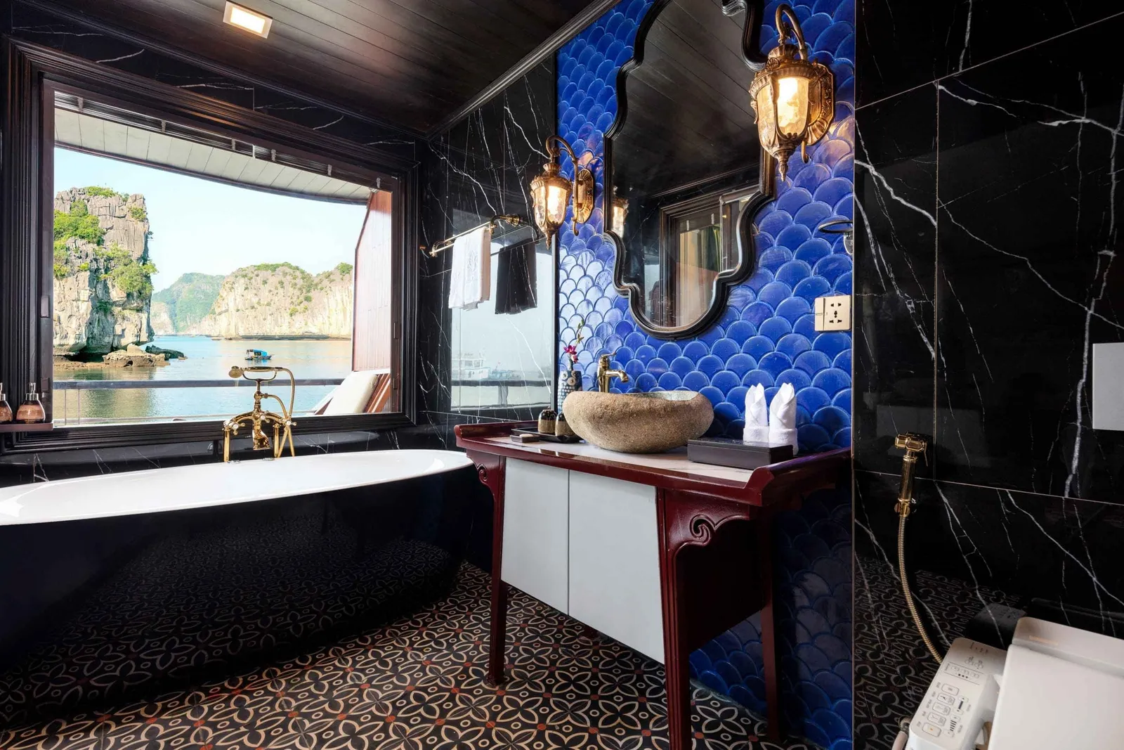 A cruise cabin bathroom with a black freestanding bathtub, blue fish-scale tiled wall, stone vessel sink and black marble walls, with a large window showing limestone karsts over calm bay water.