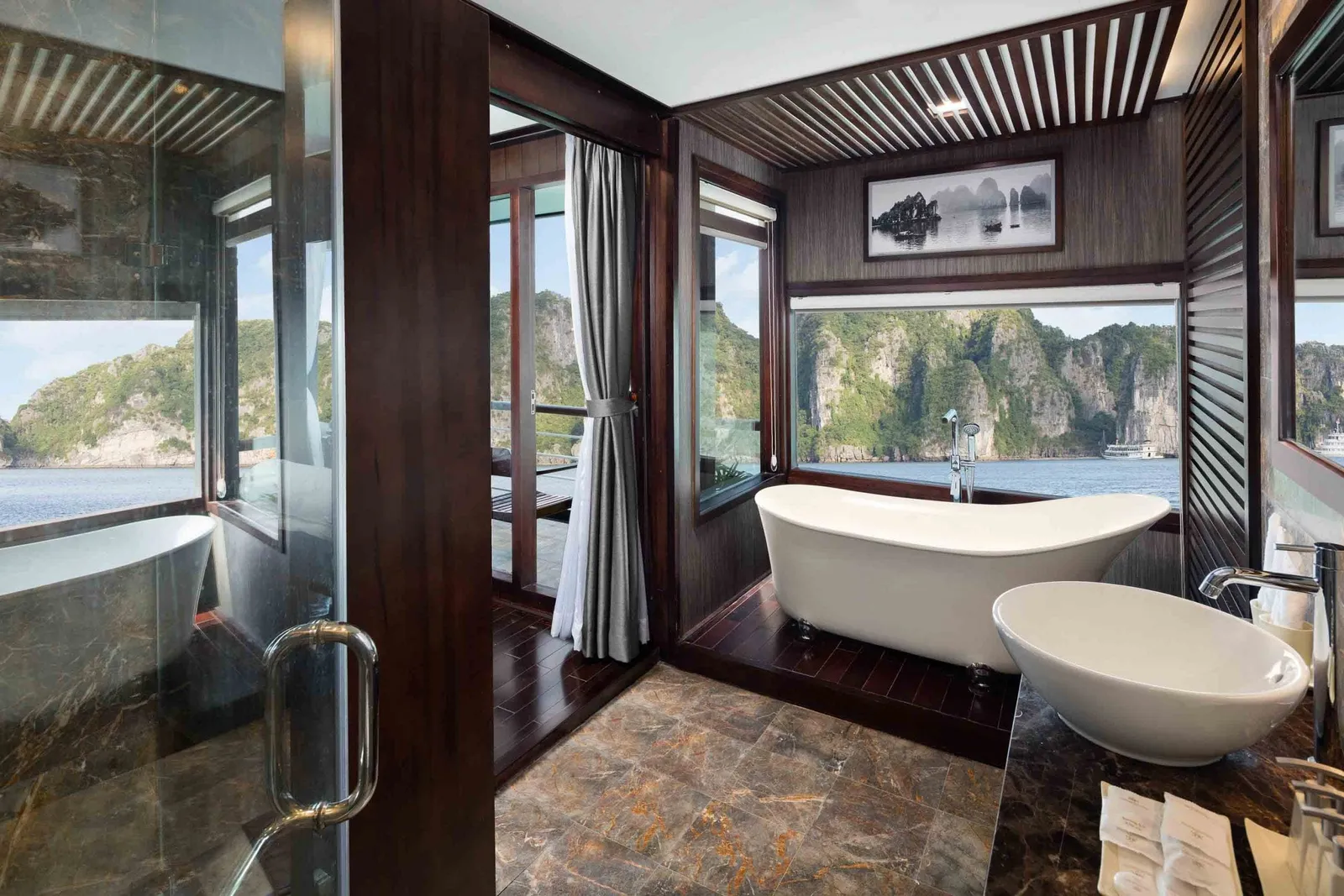 A cabin bathroom with a freestanding tub and vessel sink, dark wood and marble finishes, and large windows looking out on limestone karst islands and calm water.