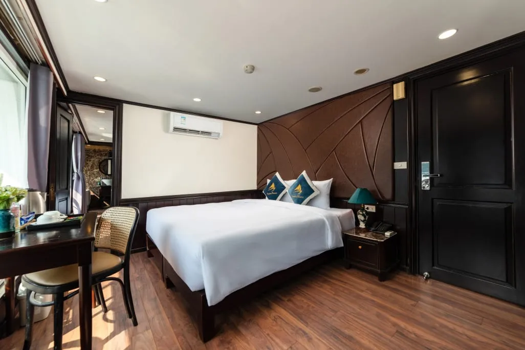 A cabin with a white-covered double bed, teal cushions bearing a sail logo, dark wood door and paneling, a desk with a chair, and a bathroom visible through an open doorway.