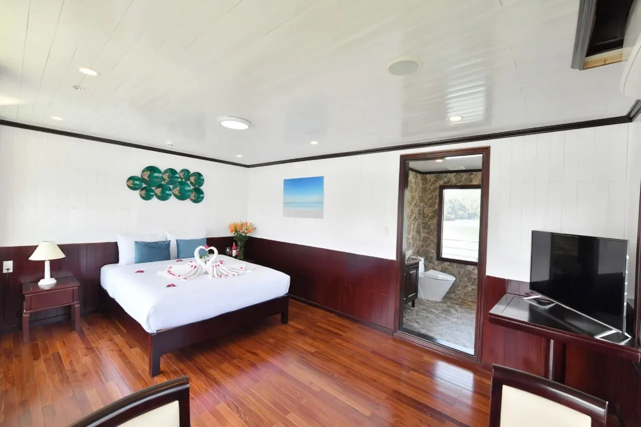 A cruise cabin with a double bed dressed in white linen with towel swans and petals, dark wood floor and paneling, a TV on the right, and an open doorway to a stone-tiled bathroom whose window shows greenery and water.