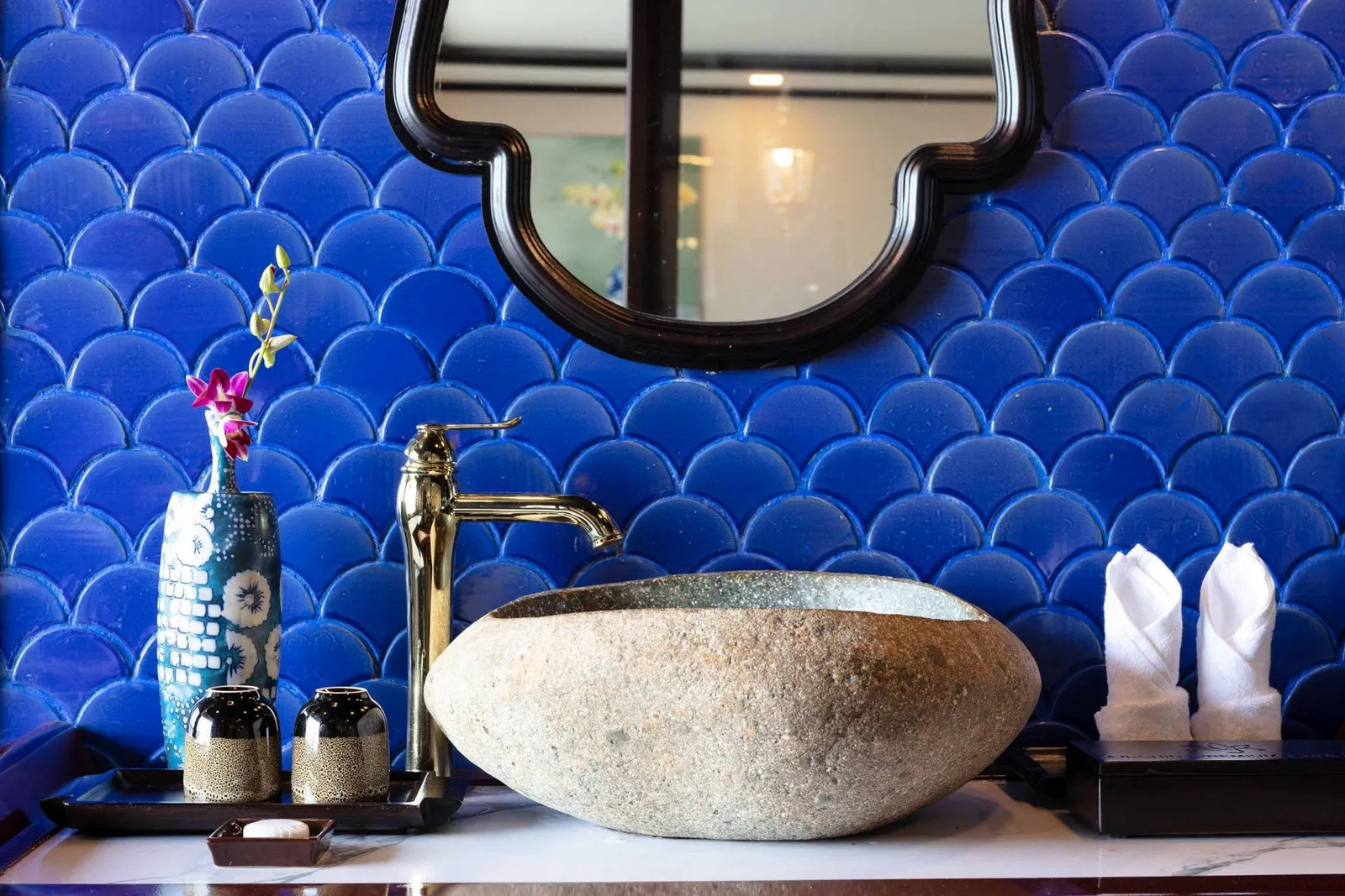 A bathroom vanity with a stone vessel sink, gold faucet, blue fish-scale tiled wall, black-framed mirror, a vase with an orchid, and folded white towels.