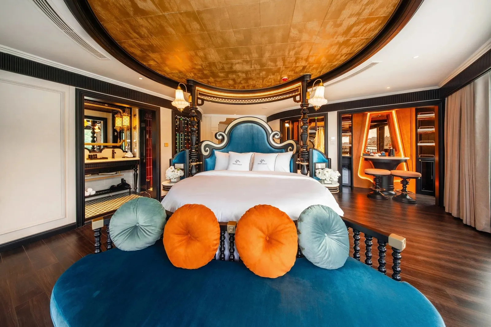 A luxurious cabin with a gold-leaf domed ceiling, a four-poster bed with blue headboard and white linens bearing a small logo on the pillows, a blue velvet bench with orange and teal round cushions, and an ensuite bathroom and bar area with stools.