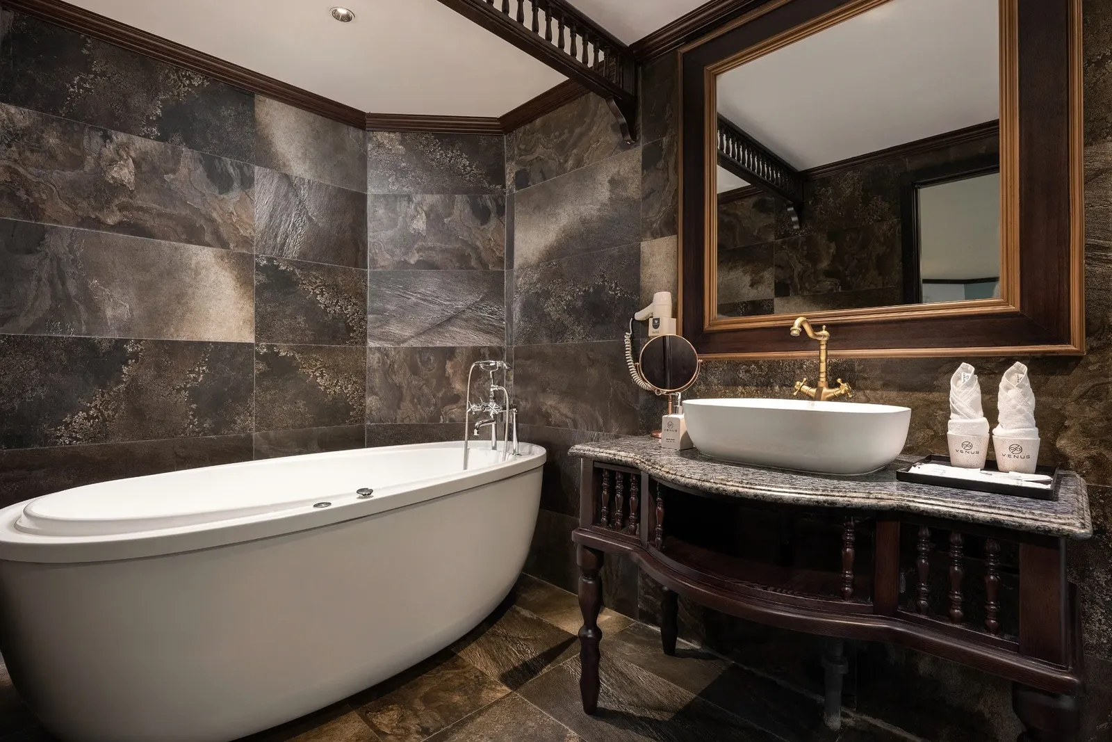 A bathroom with a white freestanding oval bathtub, dark stone-look tiles, a dark wood vanity with a white vessel sink, brass tap, framed mirror, and rolled towels on a tray bearing a 'VENUS' logo.