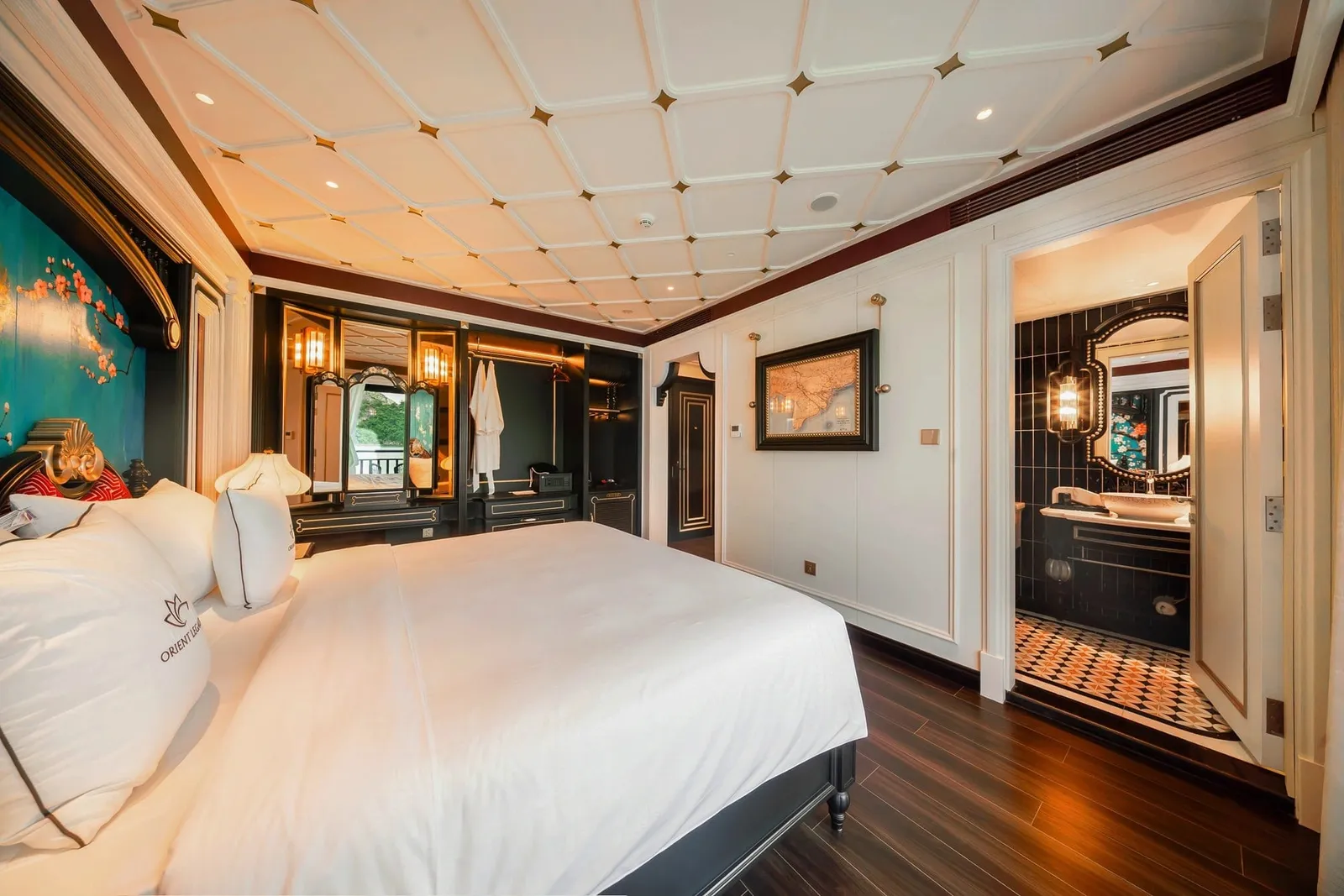 A luxurious cabin with a white-linen bed, dark wood furniture, a coffered ceiling and an open ensuite bathroom, with a lotus logo and the text 'ORIENT' embroidered on a pillow.